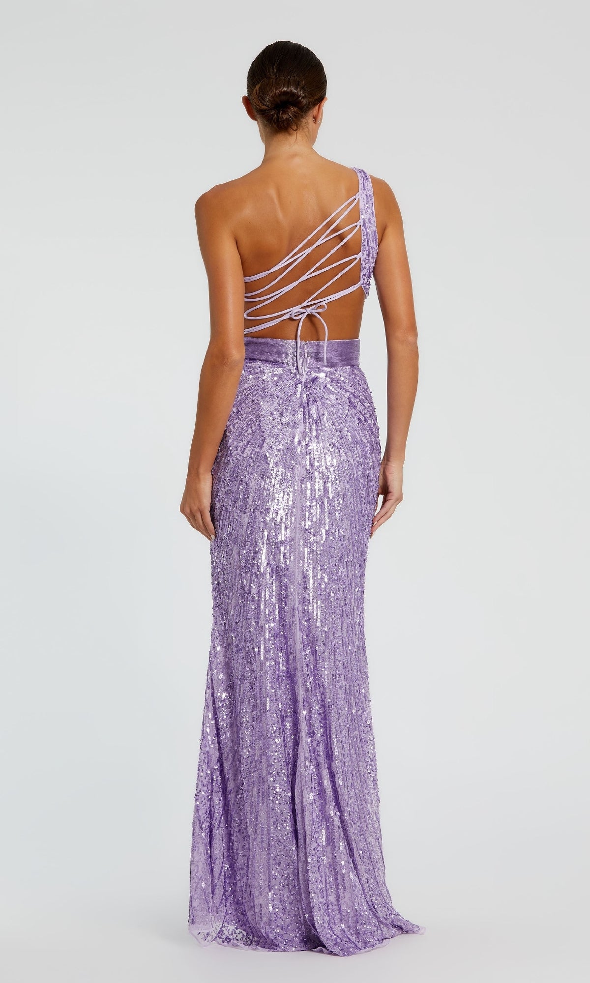 A woman stands facing away, wearing the Mac Duggal 5687 One-Shoulder Sequin Formal Dress, a floor-length lavender gown featuring an asymmetric strappy back and a fitted sequin silhouette.